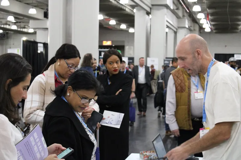Man speaking to a group of people at a trade show in a warehouse. They are looking at phones or papers.